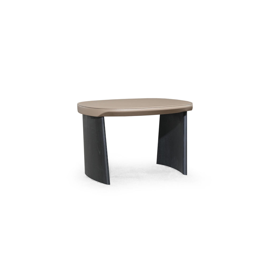 This is a low-profile stool with a rounded, flat top surface upholstered in a light beige material. It has two dark, tapered legs that curve slightly outward. The surface shows subtle stitching along the edge. Intended for seating or as a footrest, it features a minimalist design with no arms or additional hardware. Category: Furniture > Stools