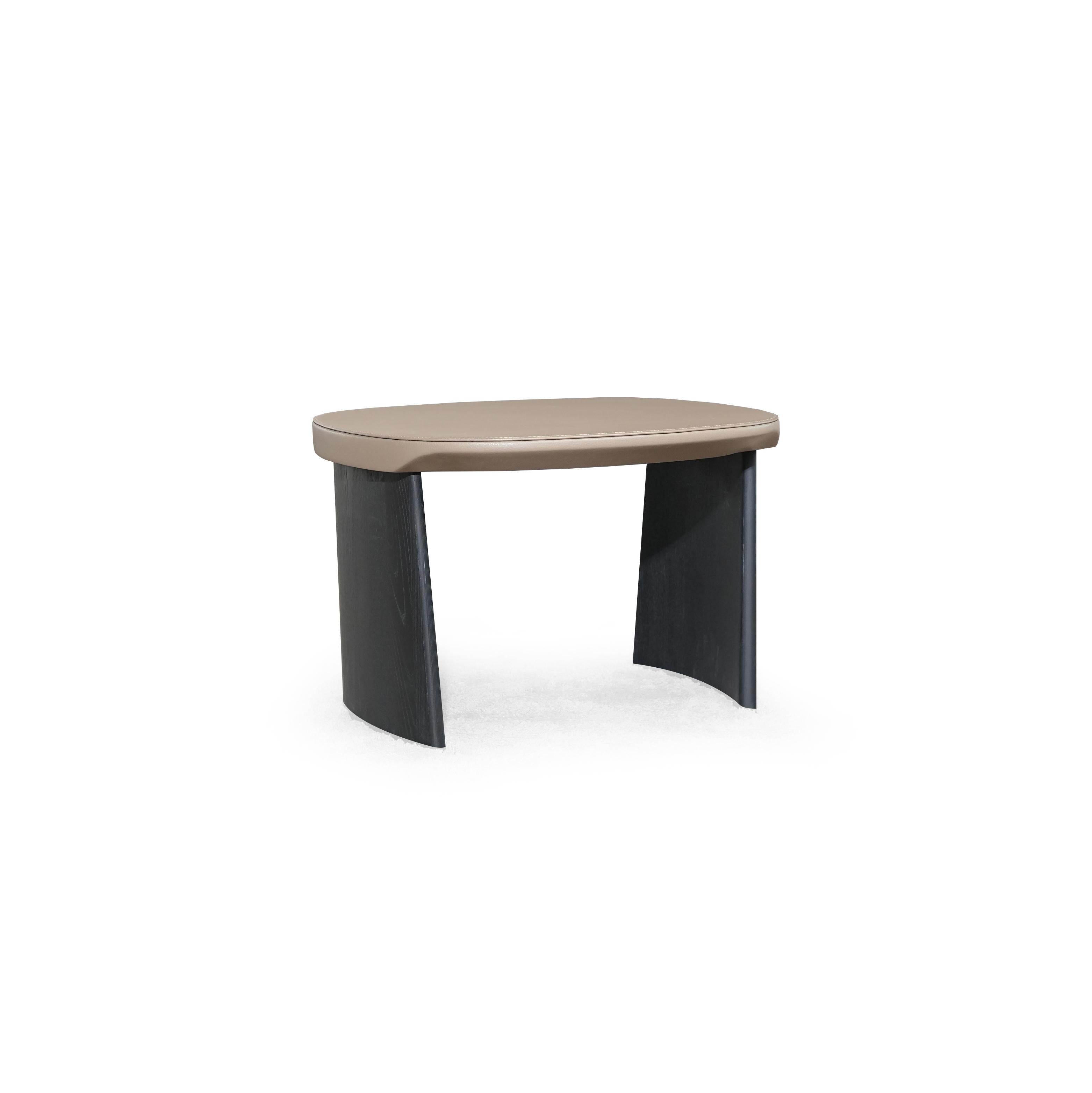 This is a low-profile stool with a rounded, flat top surface upholstered in a light beige material. It has two dark, tapered legs that curve slightly outward. The surface shows subtle stitching along the edge. Intended for seating or as a footrest, it features a minimalist design with no arms or additional hardware. Category: Furniture > Stools