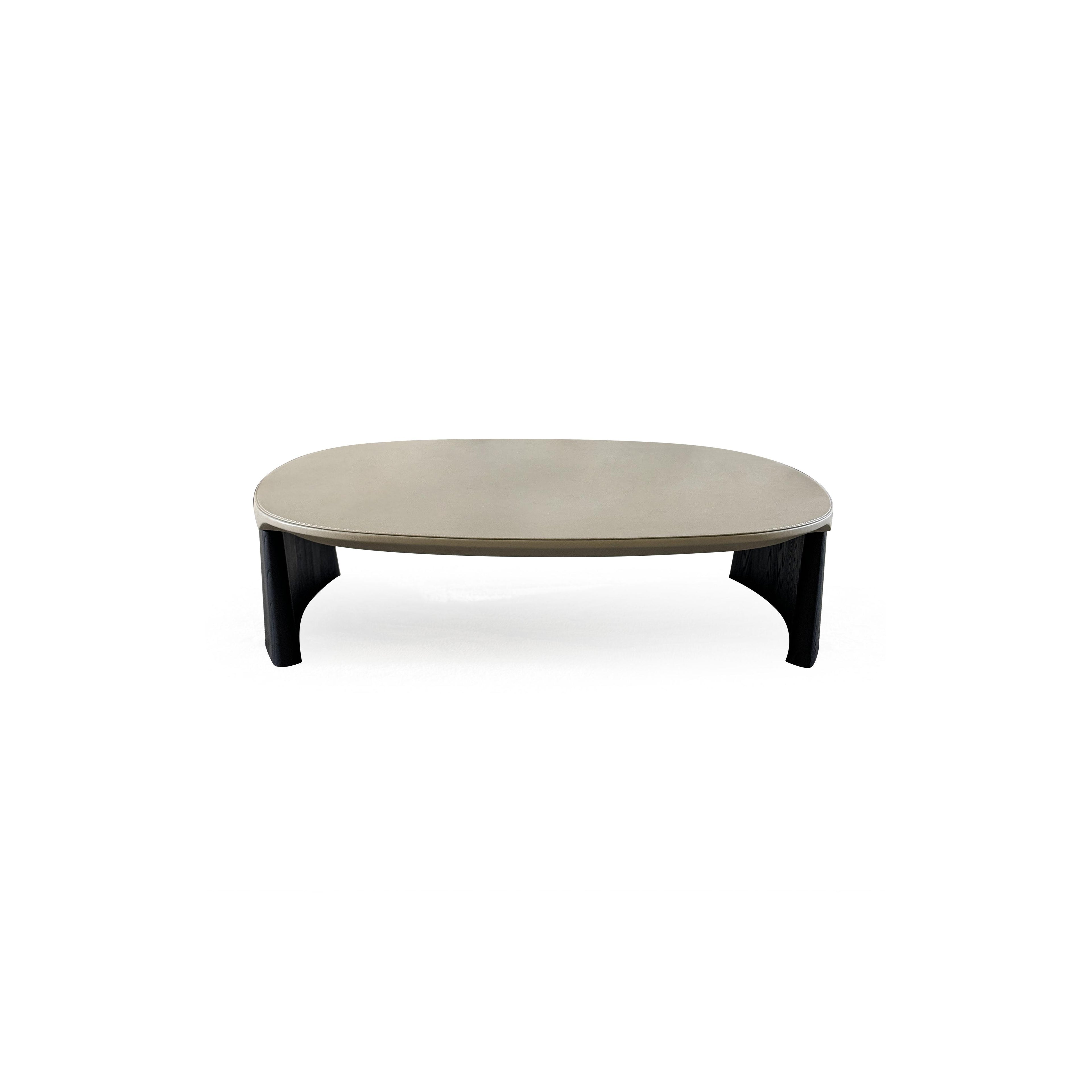 This is a low-profile stool with a rounded, flat top surface upholstered in a light beige material. It has two dark, tapered legs that curve slightly outward. The surface shows subtle stitching along the edge. Intended for seating or as a footrest, it features a minimalist design with no arms or additional hardware. Category: Furniture > Stools