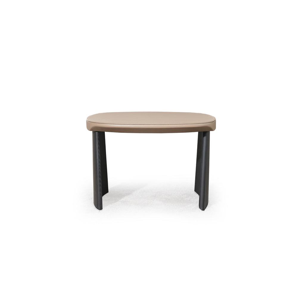 This is a low-profile stool with a rounded, flat top surface upholstered in a light beige material. It has two dark, tapered legs that curve slightly outward. The surface shows subtle stitching along the edge. Intended for seating or as a footrest, it features a minimalist design with no arms or additional hardware. Category: Furniture > Stools