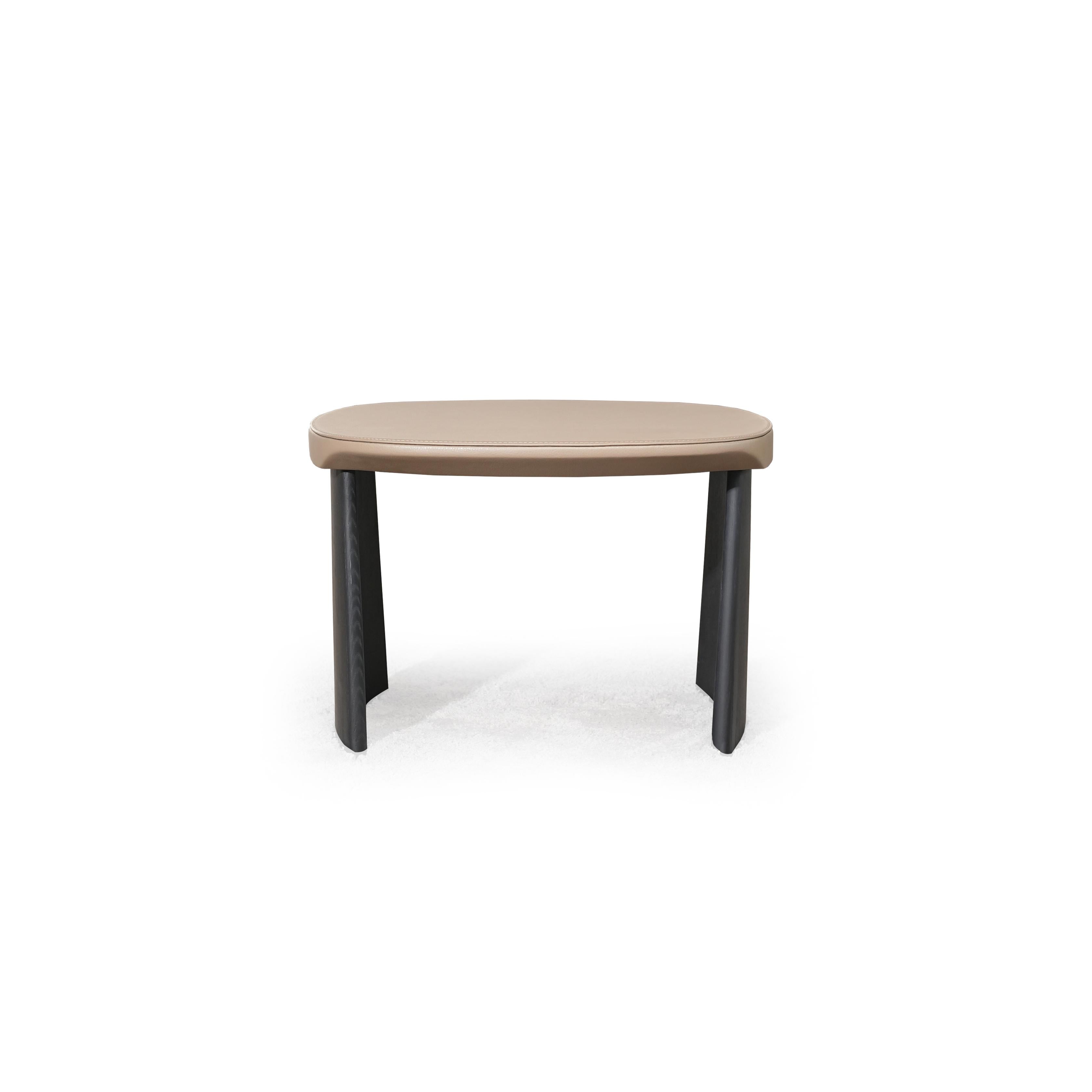 This is a low-profile stool with a rounded, flat top surface upholstered in a light beige material. It has two dark, tapered legs that curve slightly outward. The surface shows subtle stitching along the edge. Intended for seating or as a footrest, it features a minimalist design with no arms or additional hardware. Category: Furniture > Stools