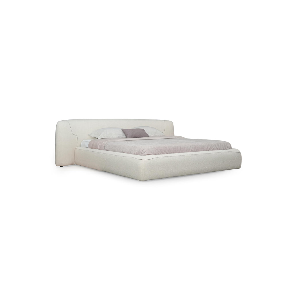 Upholstered bed with a low-profile, rectangular frame and a padded headboard featuring subtle stitched seams. Covered in a light beige fabric, it includes two white pillows and one gray accent pillow. Designed for sleeping, it has no visible legs or hardware. Category: furniture > beds > upholstered beds