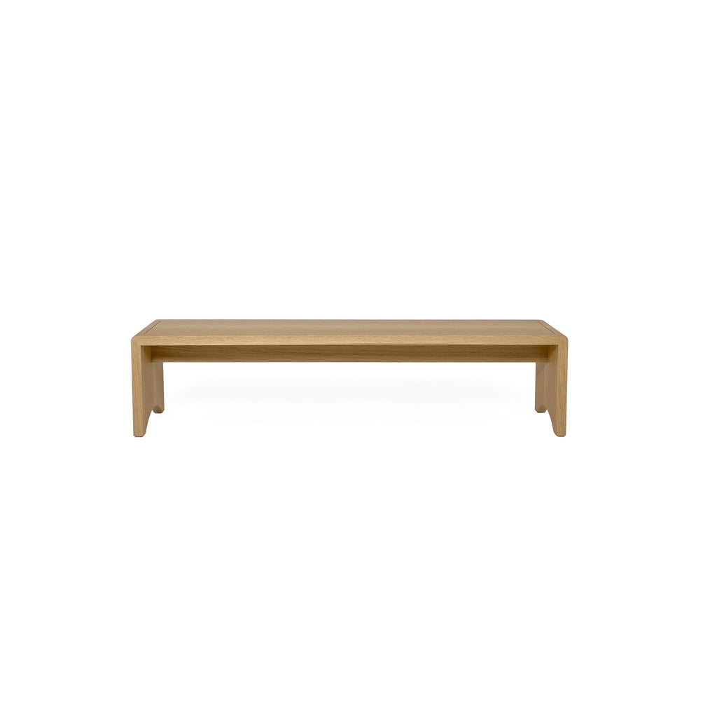 A wooden bench with a tufted beige cushion sits on four angled legs. The frame is made of light-toned wood with visible grain. The cushion has stitched seams and is held by two leather-like straps. Intended for seating, it features a simple, elongated rectangular form. Category: Furniture > Benches > Seating