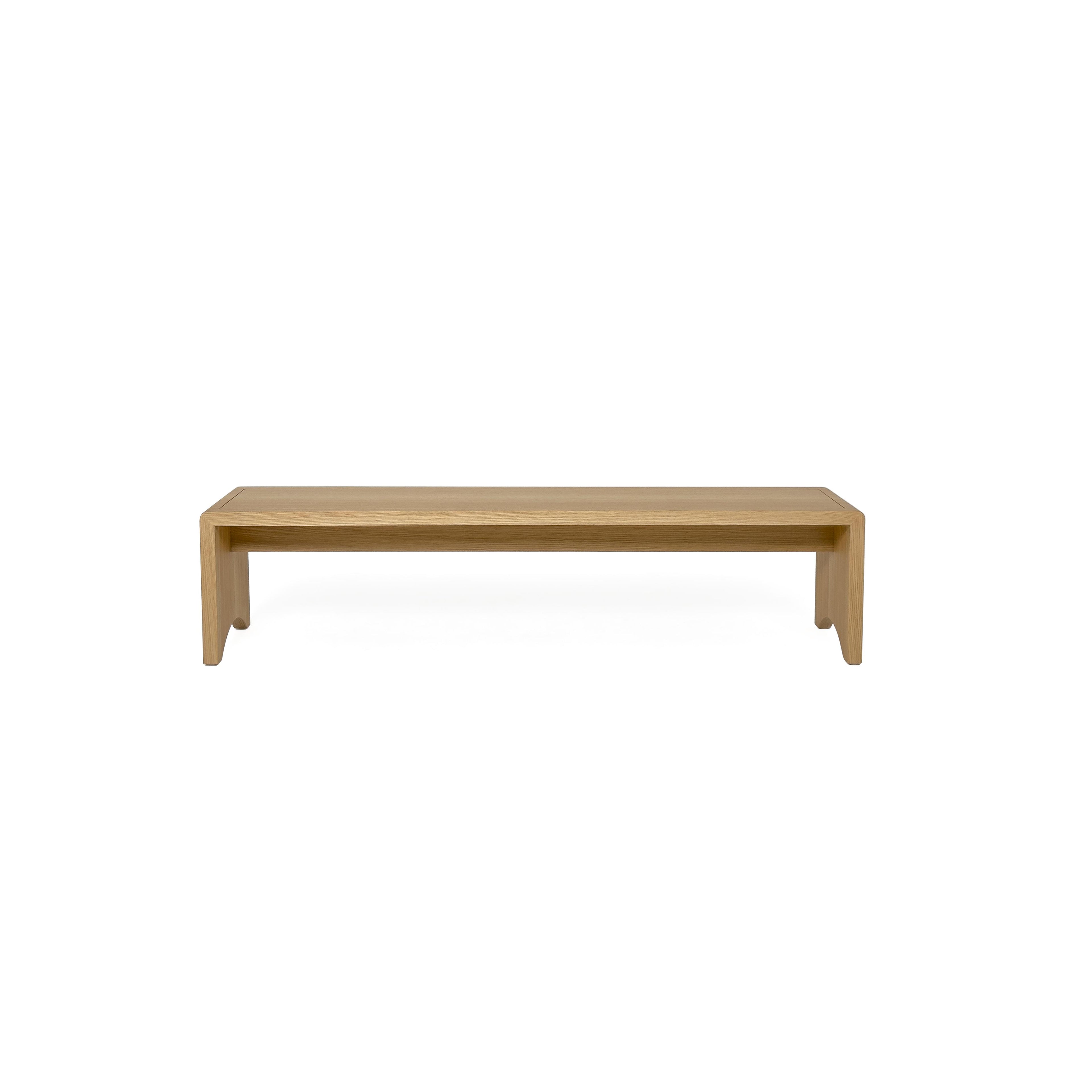 A wooden bench with a tufted beige cushion sits on four angled legs. The frame is made of light-toned wood with visible grain. The cushion has stitched seams and is held by two leather-like straps. Intended for seating, it features a simple, elongated rectangular form. Category: Furniture > Benches > Seating