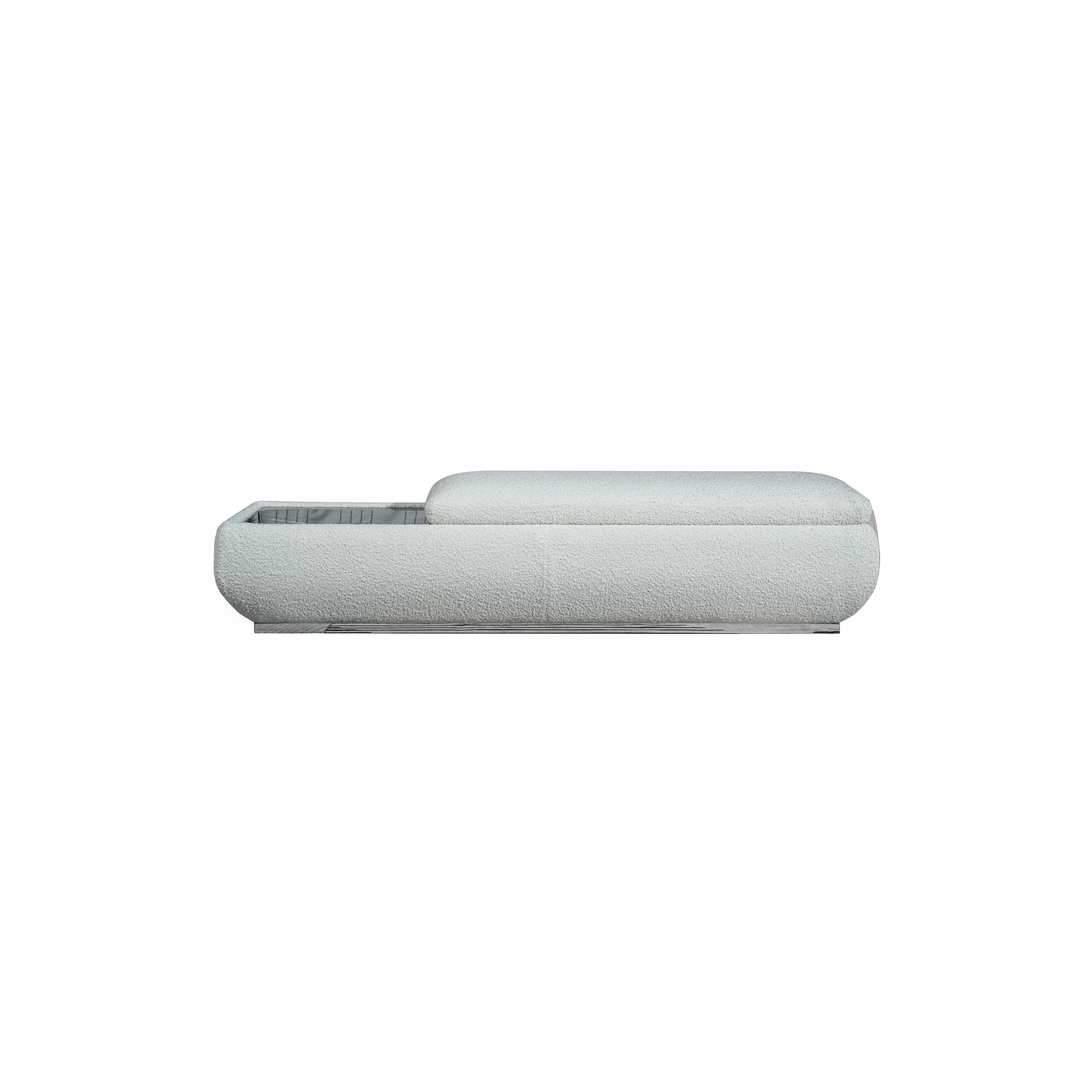 This is a low-profile bench with a soft, textured fabric upholstery in light gray. It features a rectangular base with a slightly raised cushioned section on the right. The left side reveals an open storage compartment with a grid pattern. Designed for seating and storage, it has no visible legs or frame. Category: Furniture > Seating > Bench