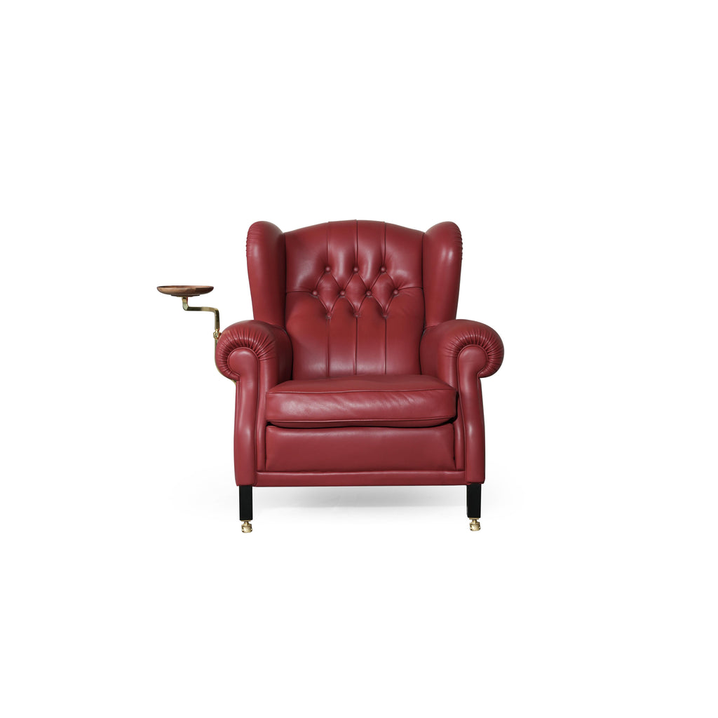 Red leather-like upholstered wingback armchair with tufted back, rolled arms, and a side-mounted wooden tray on brass armrest. Black legs with brass casters. Designed for seated comfort and display. Category: Furniture > Chairs > Armchairs