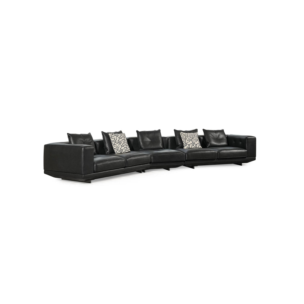 Black leather-like sectional sofa with L-shaped configuration, featuring padded backrests, armrests, and multiple cushions including one with black-and-white geometric pattern and another with zebra print. Low-profile legs support the structure. Intended for seating and lounging. Category: furniture/sofas