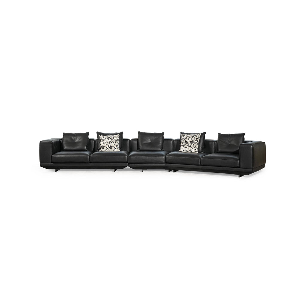 Black leather-like sectional sofa with L-shaped configuration, featuring padded backrests, armrests, and multiple cushions including one with black-and-white geometric pattern and another with zebra print. Low-profile legs support the structure. Intended for seating and lounging. Category: furniture/sofas