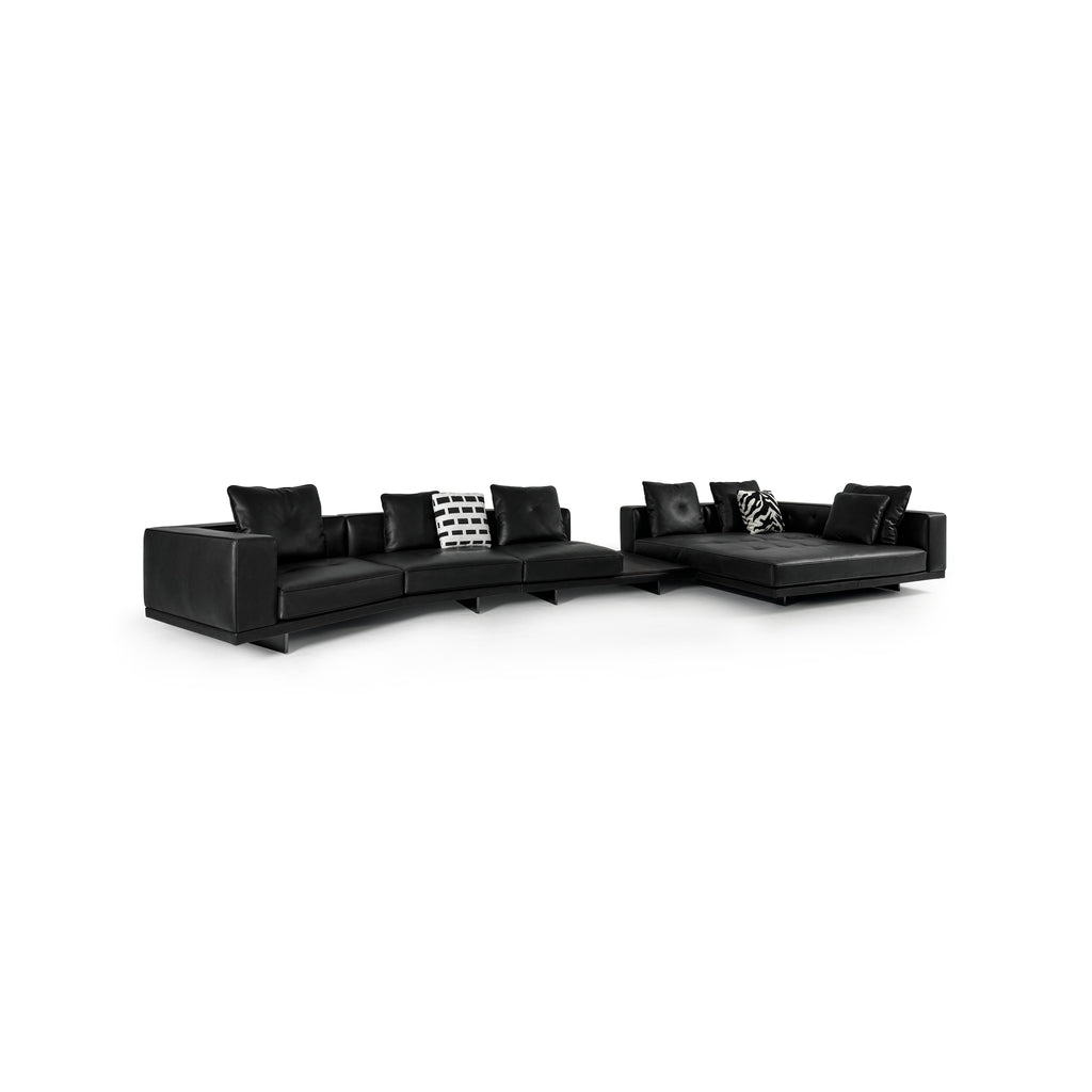 Black leather-like sectional sofa with L-shaped configuration, featuring padded backrests, armrests, and multiple cushions including one with black-and-white geometric pattern and another with zebra print. Low-profile legs support the structure. Intended for seating and lounging. Category: furniture/sofas