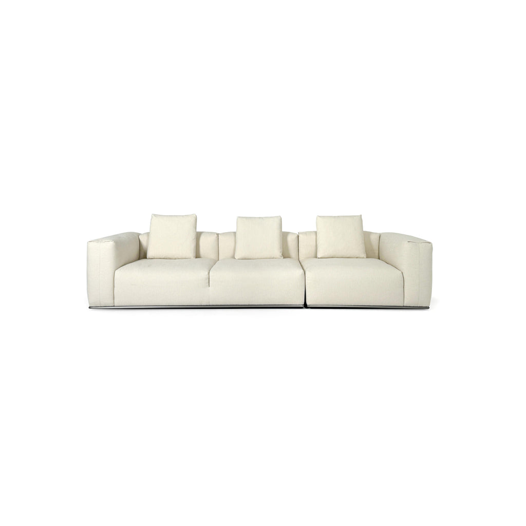 A three-seater sofa with off-white fabric upholstery, featuring segmented cushions, squared arms, and a low-profile metal base. It includes three matching back cushions. Designed for seating, it has a clean, modular form with visible stitching and no visible fasteners or handles. Category: Furniture > Sofas > Sectionals