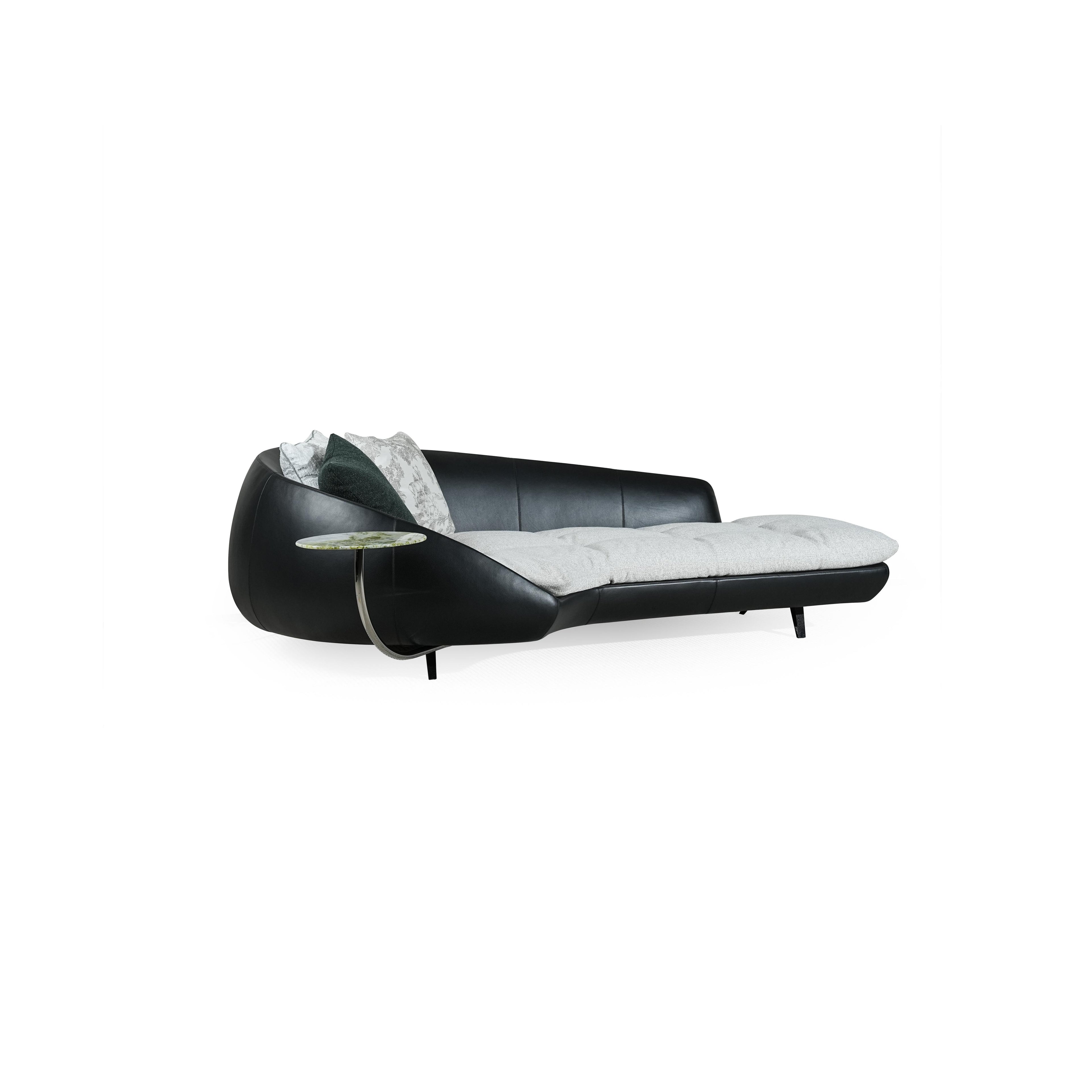 Modern chaise lounge with black leather-like frame and light gray cushioned seat. Curved backrest, single armrest with circular glass table, and four short legs. Two pillows: one dark green, one patterned. Designed for lounging or reclining. Category: Furniture > Sofas & Chaises
