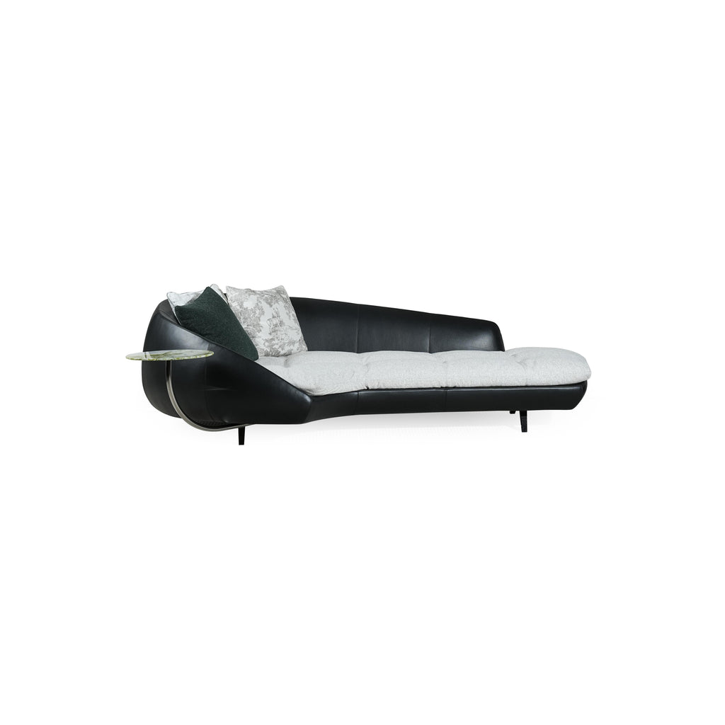Modern chaise lounge with black leather-like frame and light gray cushioned seat. Curved backrest, single armrest with circular glass table, and four short legs. Two pillows: one dark green, one patterned. Designed for lounging or reclining. Category: Furniture > Sofas & Chaises