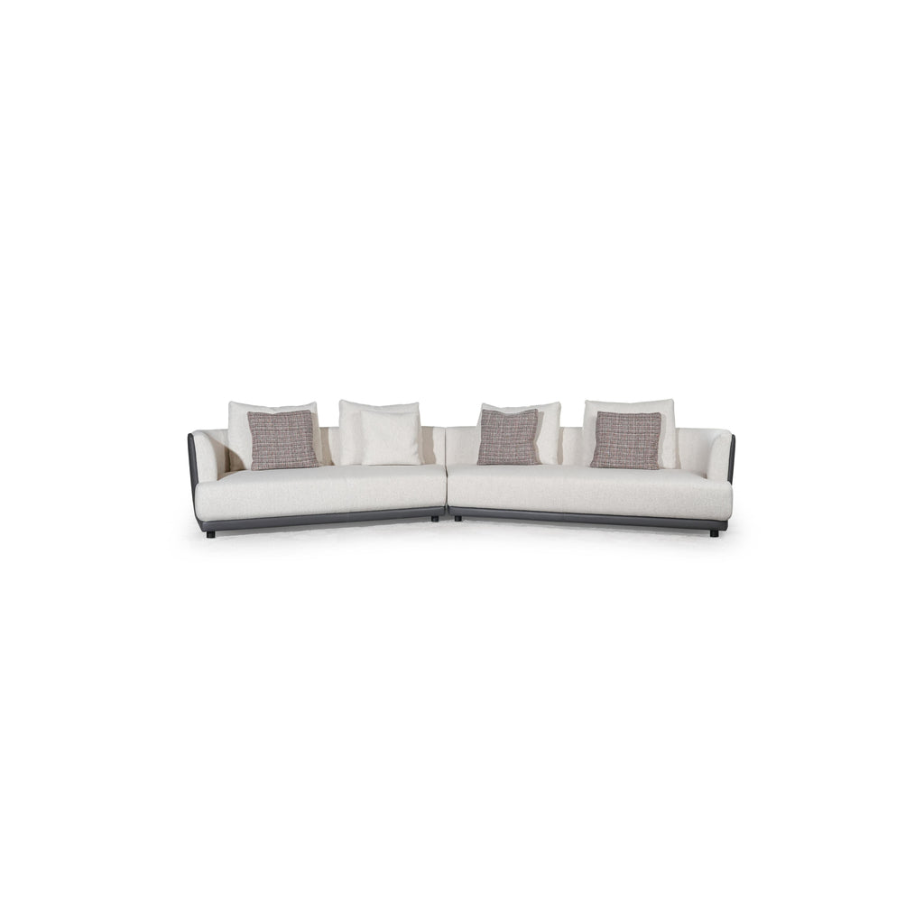 L-shaped sectional sofa with off-white fabric upholstery and dark gray armrests and base. Features multiple cushions, including two textured throw pillows. Supported by short, dark legs. Designed for seating and lounging. Category: furniture/sofas