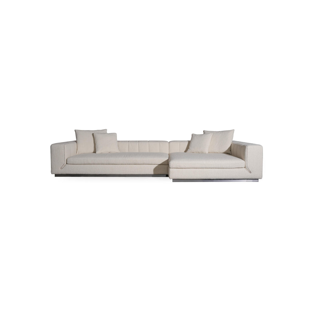 L-shaped sectional sofa with off-white textured fabric upholstery, featuring padded backrest, cushioned arms, and low-profile metal base. Includes multiple throw pillows. Designed for seating and lounging. Category: furniture/sofas
