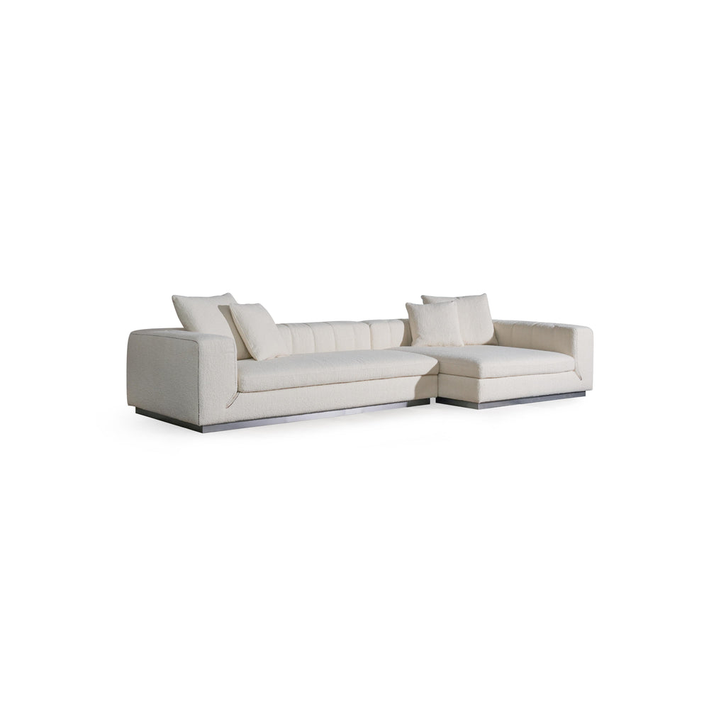 L-shaped sectional sofa with off-white textured fabric upholstery, featuring padded backrest, cushioned arms, and low-profile metal base. Includes multiple throw pillows. Designed for seating and lounging. Category: furniture/sofas