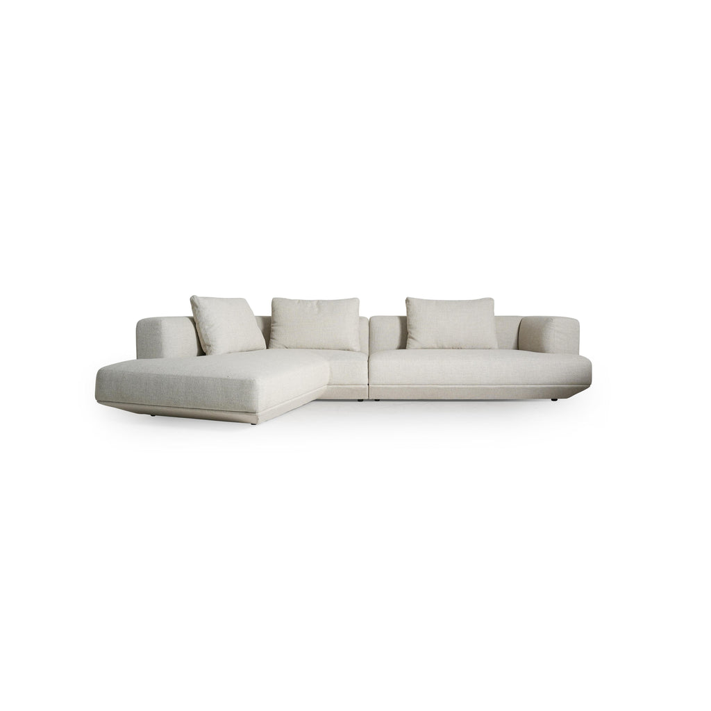 This is a modular sectional sofa with a light beige fabric upholstery, featuring three cushions, two armrests, and a chaise lounge on the left. It has low-profile legs and a clean, rectangular form. Designed for seating, it includes visible stitching along seams and a consistent fabric texture. Category: furniture > sofas > sectional sofas